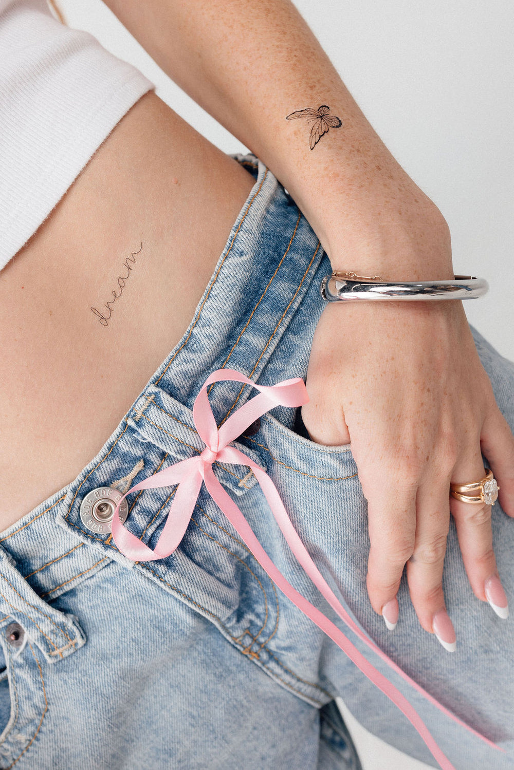 Close-up of fine-line butterfly and script hip tattoos above light-wash jeans tied with a pink ribbon